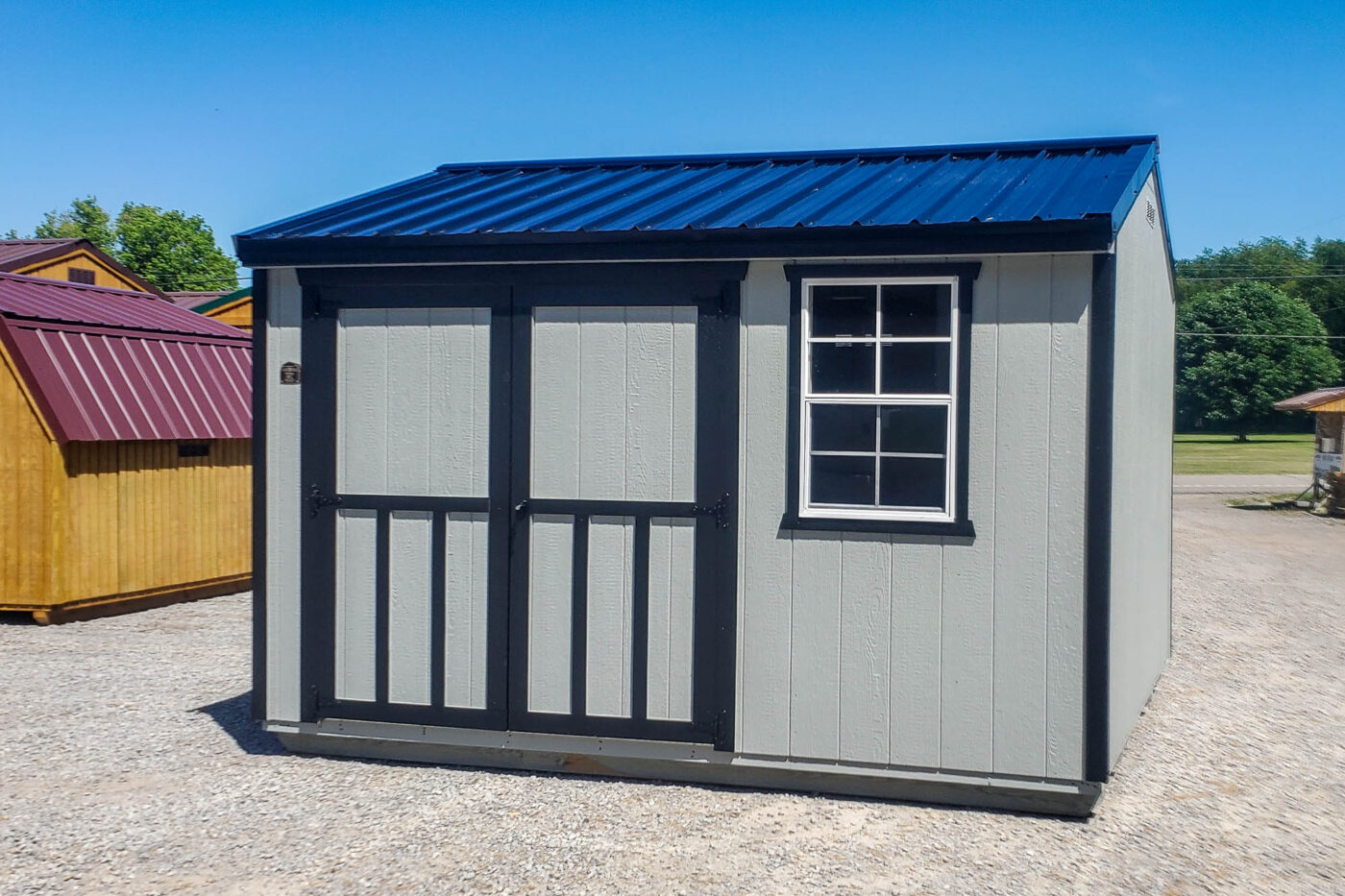 Front and left-side view of a 10x12 gray utility shed with black trim and a blue metal gable roof. Features a single window and a double door with transom windows.