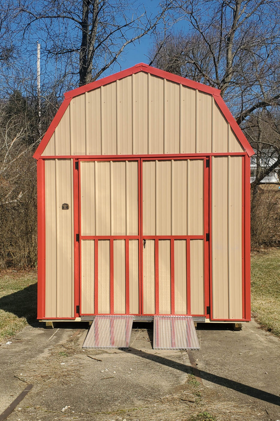 A front view of a beige metal barn shed with red trim and a sliding door, positioned on a wooden base with a ramp leading to the entrance.