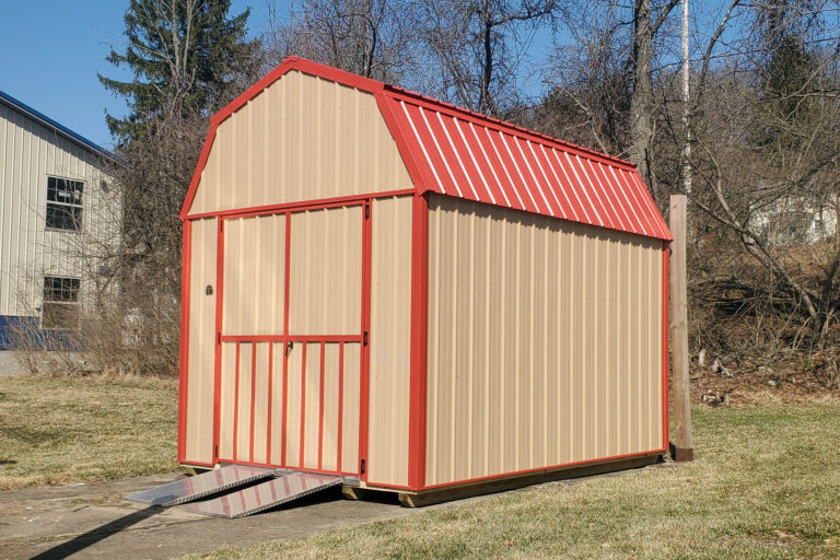 A beige metal shed with red trim and a red barn-style roof, featuring a single door and a wooden ramp at the entrance. The shed sits on a grassy lot with a house and trees in the background.