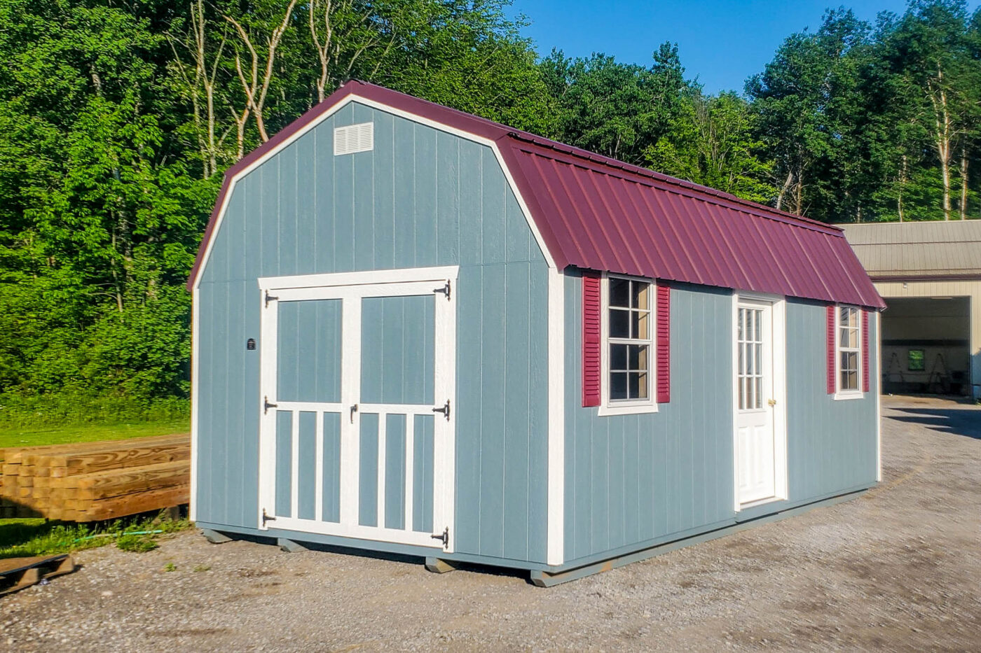 Light blue barn-style shed with maroon metal roof, double barn doors with windows, and white trim, situated on paved ground.