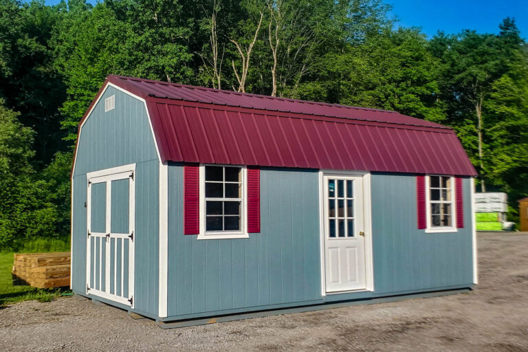 Light blue barn-style portable building with maroon metal roof, three windows, and a white door, displayed on a gravel lot.