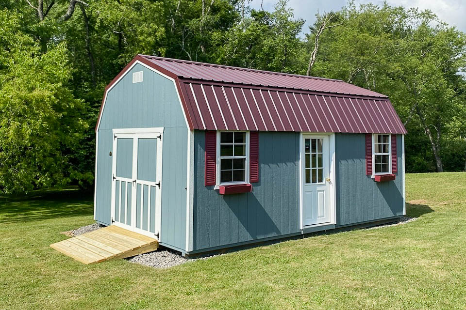 Light blue barn-style shed with maroon metal roof, two front windows with decorative shutters, double door entrance, and a wooden ramp, situated on a grassy lawn.