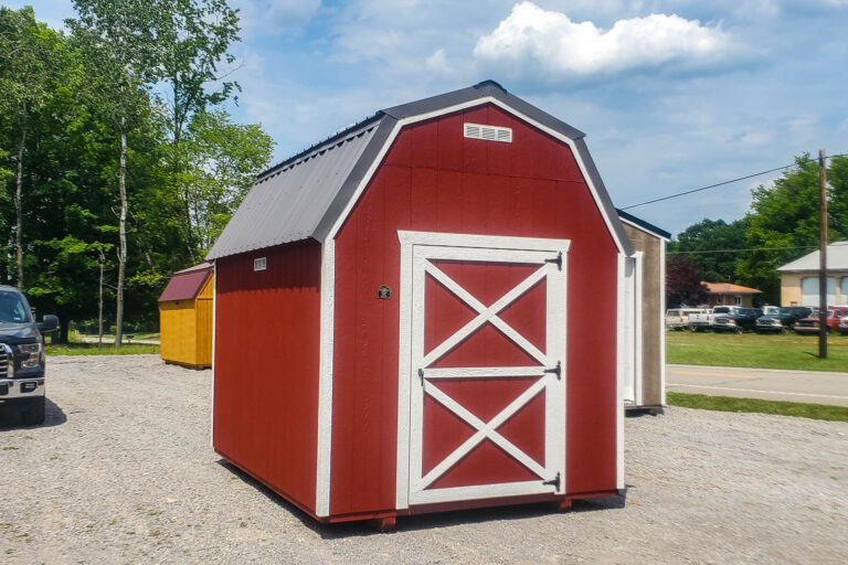 A small red barn-style shed with white trim and a gambrel gray metal roof, featuring a single white-trimmed door with crossbuck design, sitting on gravel.