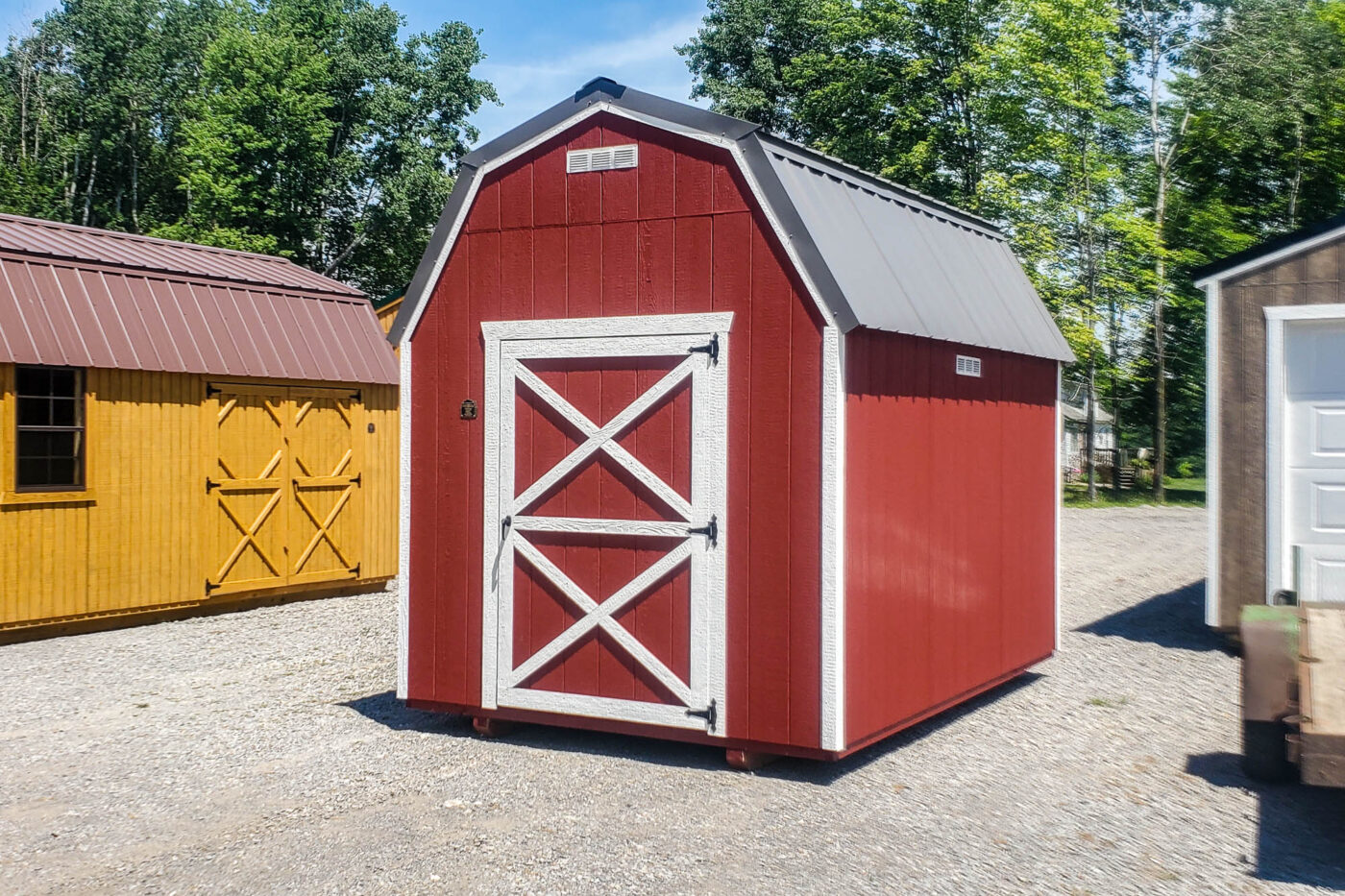 An 8x10 red barn-style shed with a gray gambrel roof and white trim, placed on gravel near other wooden buildings, under a sunny sky.