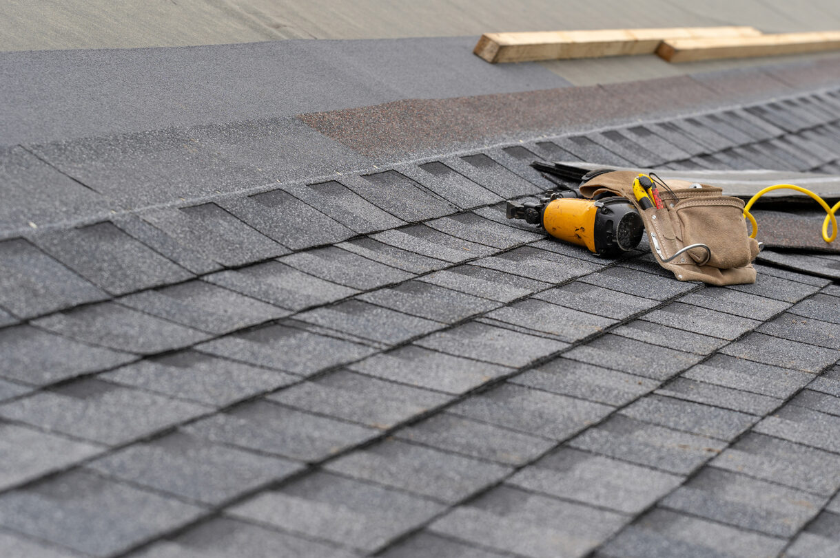 photo of toolbelt with instrument and nail gun lying on asphalt or bitumen shingle on top of the new roof under construction residential house or building