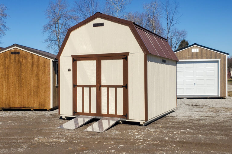 Tan and brown lofted barn on dirt and gravel lot - Tan wood siding with brown trim on corners and double doors beneath brown metal roof