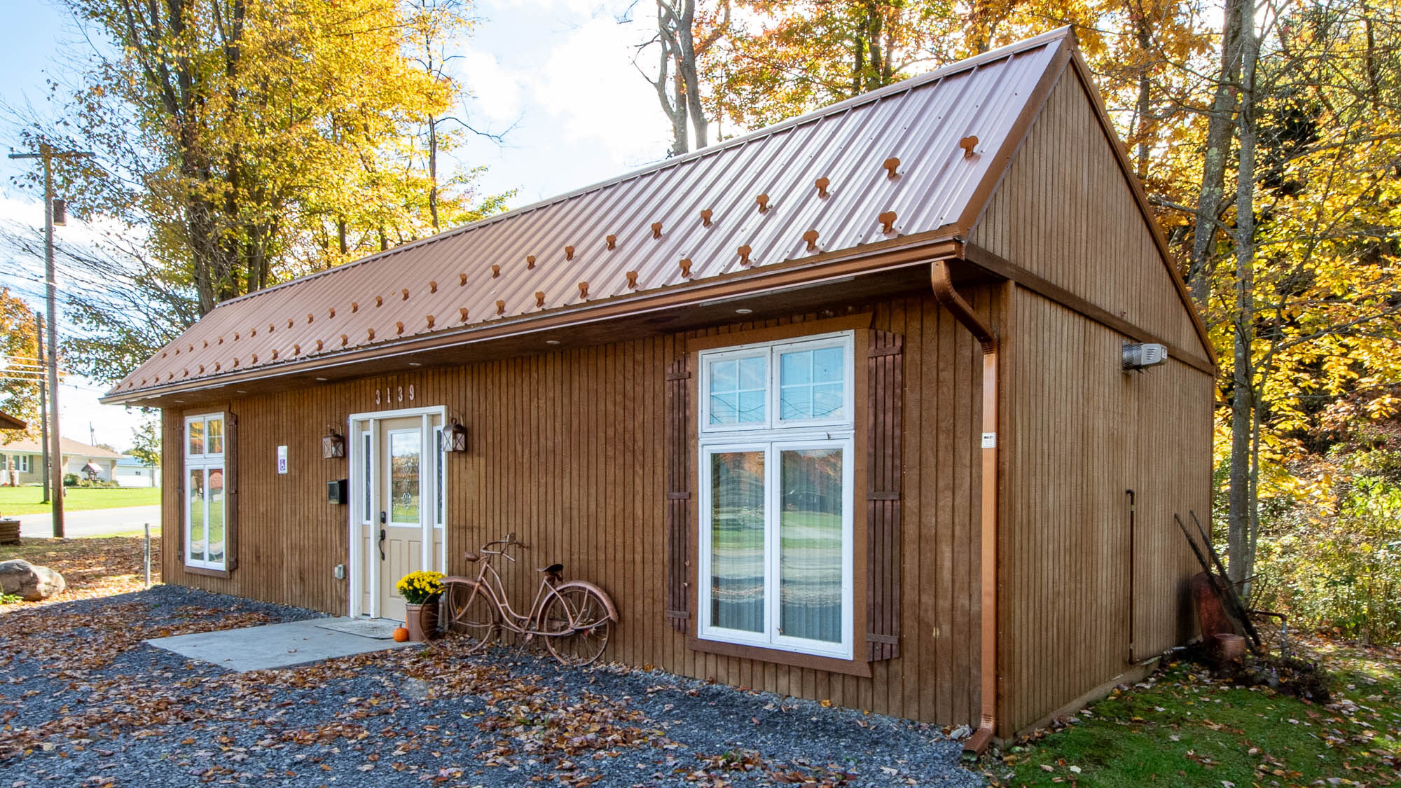 Brown single-story building with metal roof, large front windows, and a white door, set among autumn trees with a bicycle and potted flowers at the entrance.