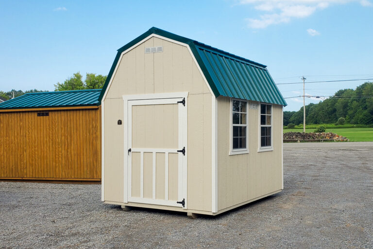 Light beige and green lofted barn on gravel lot - Light beige painted wood shed with white trimmed corners windows and door under green metal roof - trees in the distance