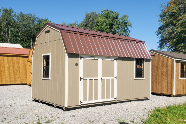 Tan lofted barn with brown metal roof - tan wood siding with white trimmed corners, windows and double doors under brown metal roof - on gravel tree-lined lot among other wooden sheds in the background