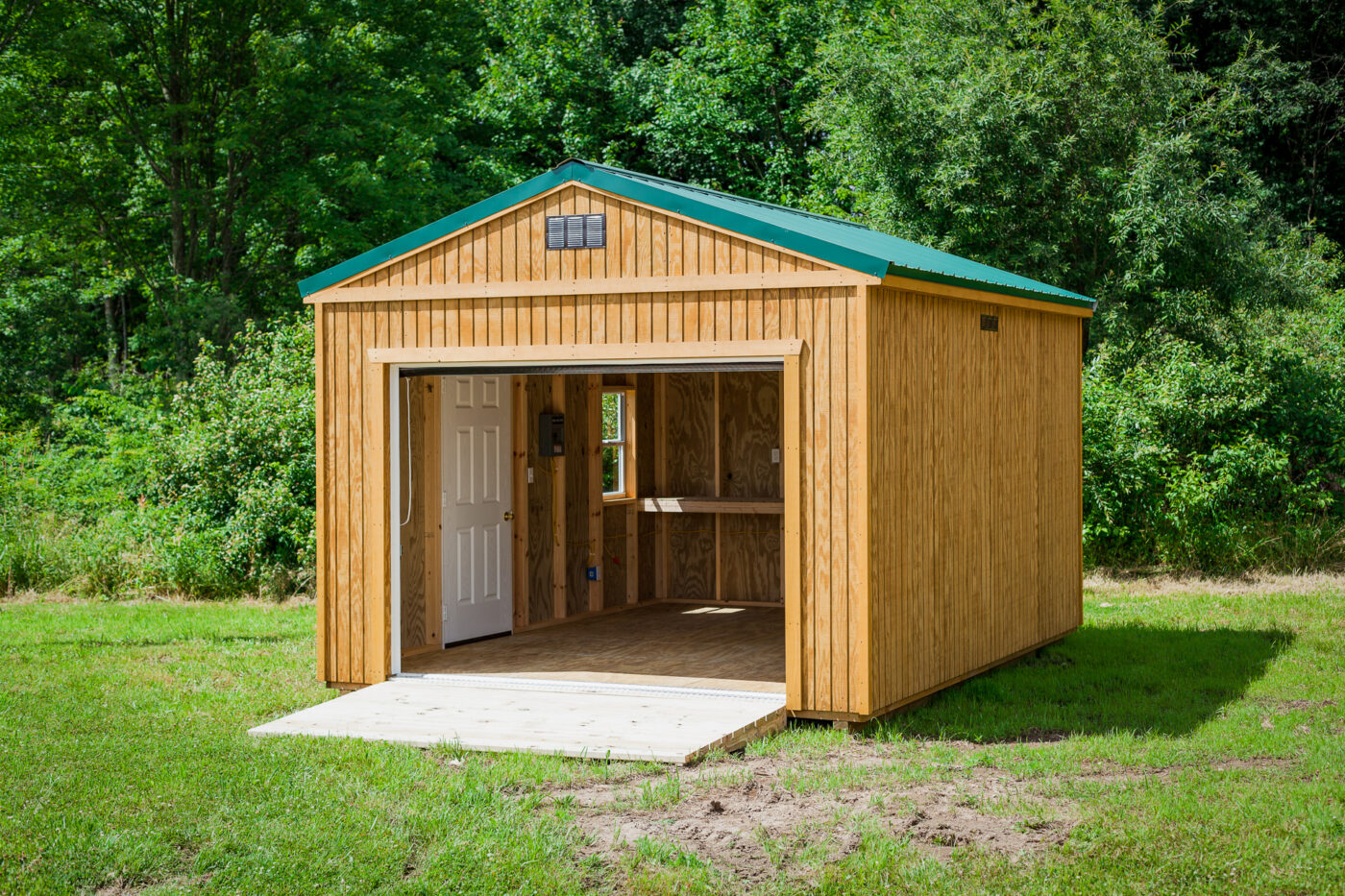 Portable garage with wood siding, green metal roof, ramp and side door sitting on a property