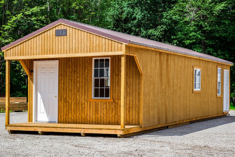 Wooden portable cabin with front porch, white entry door, and gable roof set against a wooded background.

