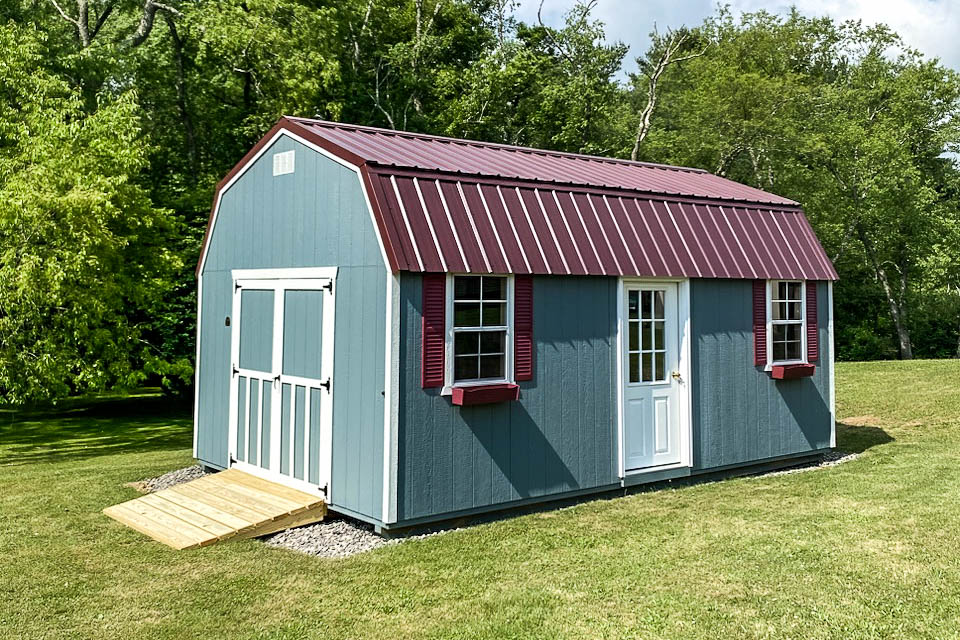 Blue lofted barn shed with maroon roof and shutters, double doors and entry door, placed on lawn near trees.