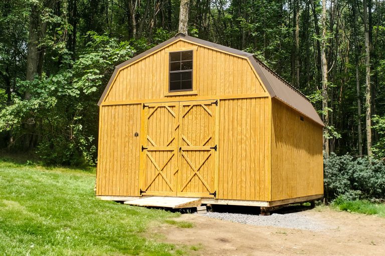 Wood lofted barn shed with double doors and gambrel roof, set near woods on a gravel base.