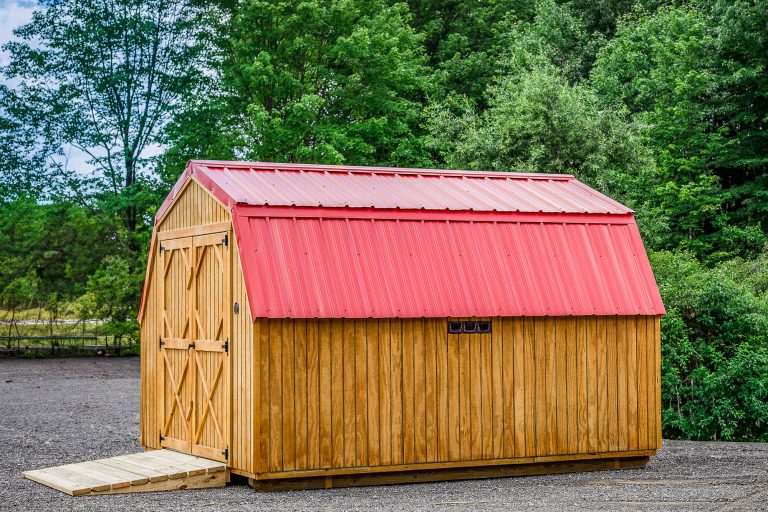 Wooden mini barn with bright red metal roof, side vent, and double doors with ramp, placed on gravel lot.