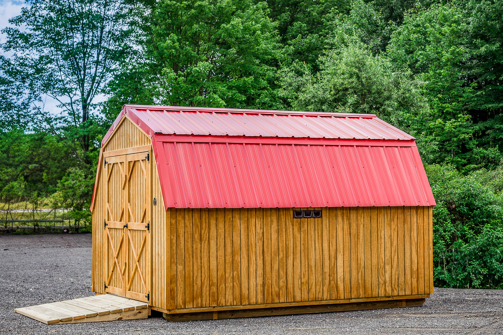 Prefab barn with bright red metal roof, side vent, and double doors with ramp, positioned on gravel lot.